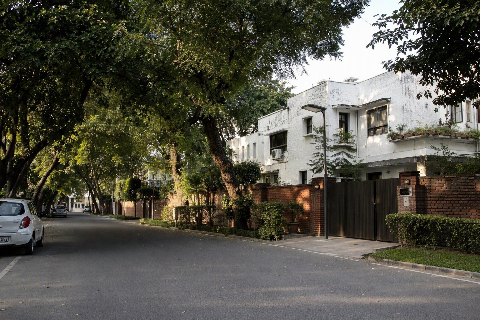 Tree lined DLF Phase 1 Gurugram street with older whitewashed bungalows behind brick boundary walls and parked cars